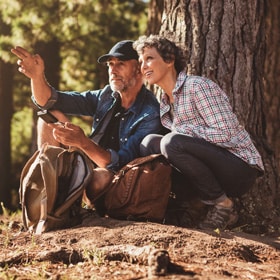 A retired couple crouches near a tree while camping